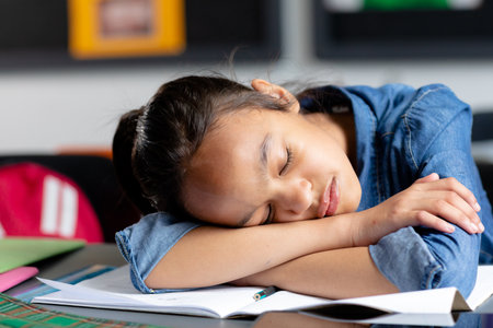 Tired biracial schoolgirl sitting at desk, asleep in class, copy space. Education, childhood, elementary school, inclusivity and learning concept.の写真素材