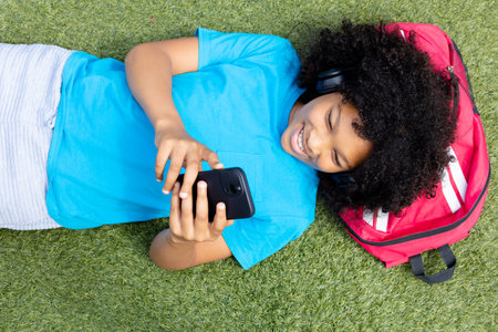 Overhead view of smiling biracial schoolgirl lying on grass using smartphone. Education, childhood,...