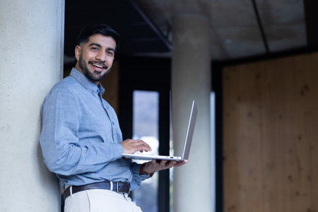 Smiling man using laptop while leaning against column in modern office, copy space. Business, technology, entrepreneur, corporate, professional, workspaceの写真素材