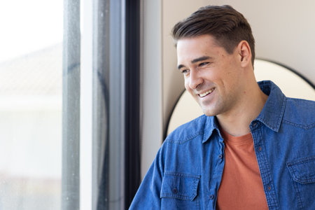 Smiling man in casual denim shirt standing by window, looking outside. Portrait, confident, happy, thoughtful, relaxed, indoorsの写真素材