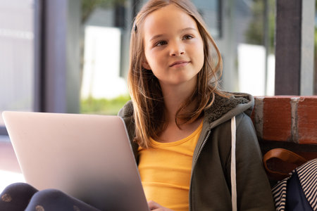 Smiling caucasian schoolgirl using laptop, sitting outside school, with copy space. Education,...