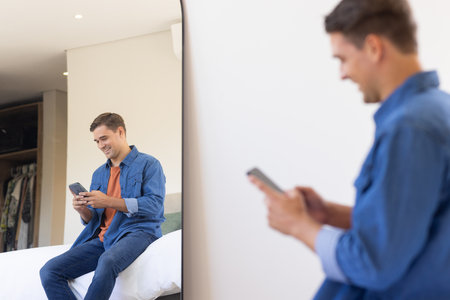 Using smartphone, man sitting on bed and smiling at reflection in mirror. Technology, bedroom, happy, morning, casual, relaxationの写真素材
