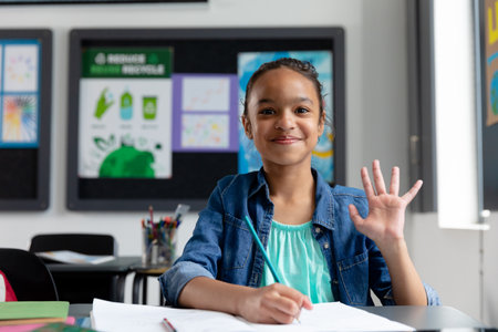 Portrait of happy biracial schoolgirl sitting at desk in class, waving, with copy space. Education,...