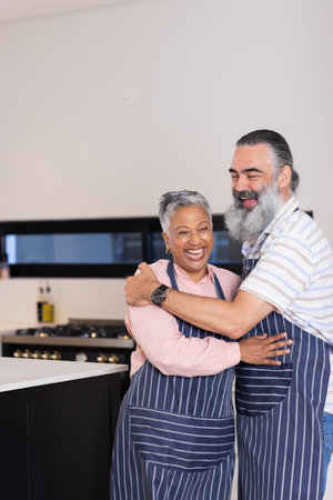 Senior couple in aprons hugging and smiling in modern kitchen. Lifestyle, cooking, happiness, love, affection, elderlyの写真素材