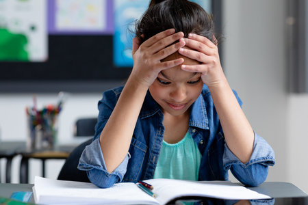 Stressed biracial schoolgirl sitting at desk in class, head in hands, staring at book, copy space. Education, childhood, elementary school, inclusivity and learning concept.の写真素材