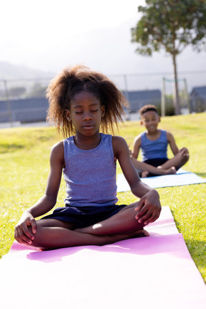 Happy african american schoolchildren doing yoga and meditating on field at school. Elementary school, sports, education and learning concept.の写真素材