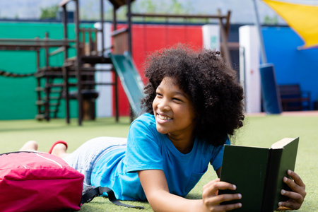 Smiling biracial schoolgirl lying in school playground with book, looking away, copy space. Education, childhood, elementary school, inclusivity and learning concept.の写真素材
