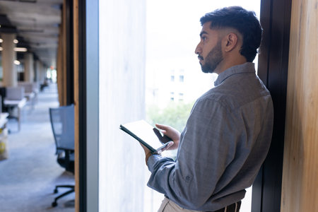 Holding tablet, man standing by window in modern office environment. Business, technology,...