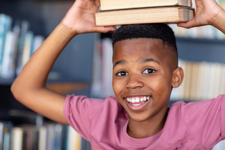 Portrait of smiling african american boy holding books on head in school library. Education, childhood, inclusivity, elementary school and learning concept.の写真素材