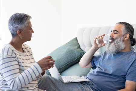Senior man drinking water while using laptop, talking with woman on bed. Elderly, hydration,...