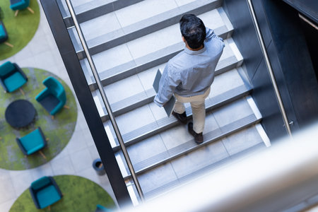 Walking down stairs, man holding laptop in modern office environment. Business, technology, professional, productivity, corporate, workplaceの写真素材