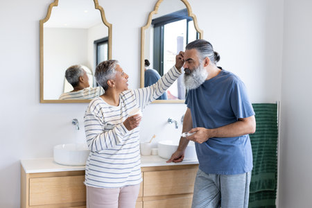 Senior couple in bathroom, woman applying cream to man's face, smiling together. skincare, happiness, wellness, routine, elderly, beautyの写真素材