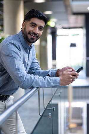 Smiling businessman holding smartphone, leaning on railing in modern office. entrepreneur,...