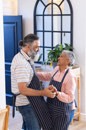 Senior couple dancing together in kitchen, wearing aprons and smiling joyfully. Seniors, happiness, lifestyle, relationship, activity, funの写真素材