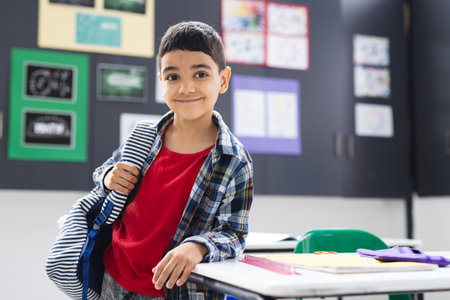 In school, young biracial boy with a backpack stands smiling in the classroom, copy space. He has dark hair, brown eyes, and wears a plaid shirt over a red tee, unalteredの写真素材