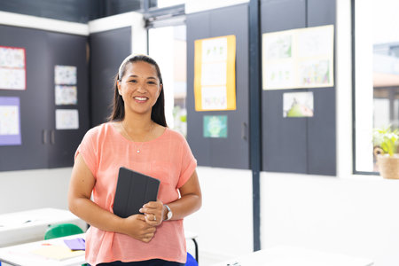 In school, young biracial female teacher holding a tablet, smiling, with copy space, in classroom....