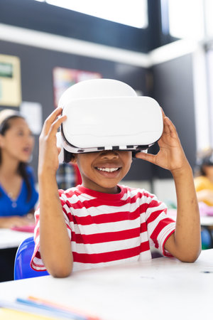 In school, young African American boy wearing a VR headset smiles in the classroom. Behind him, a biracial woman and children focus on their tasks, unaltered.の写真素材