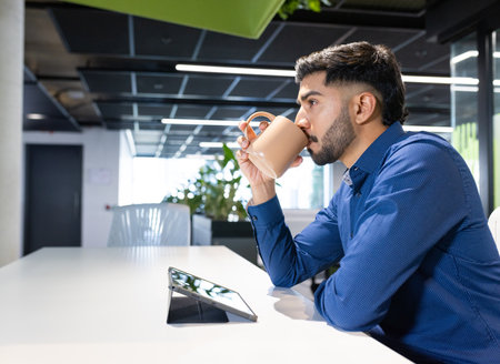 Using tablet, man drinking coffee in modern office workspace, copy space. Business, technology, corporate, productivity, professional, workingの写真素材