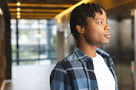 African American man wearing plaid shirt standing indoors in a modern business office with copy space. He has short twisted hair, looking away thoughtfully, unaltered.の写真素材