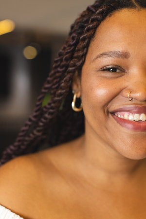 A young biracial woman is smiling, wearing nose ring and hoop earrings, in a modern business office. She has brown eyes, braided hair, and a warm complexion, unaltered.の写真素材