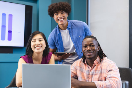 Diverse group of young colleagues working together in a modern business office, smiling at camera. Asian woman holding tablet, biracial and African American men with laptops, unalteredの写真素材