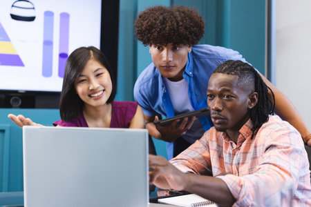 Diverse group of young colleagues focusing on laptop screen in a modern business office. Asian female with long black hair holds tablet, African American and biracial males wear casual shirts, unalteredの写真素材