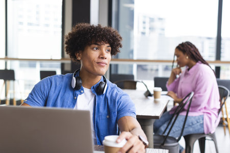 Young biracial man with curly hair working on laptop in a modern business office, woman in background. He has headphones, wearing a blue shirt, she has a pink top, both holding coffee cups, unaltered.の写真素材