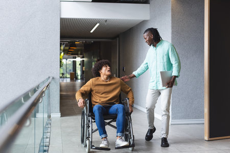 African American man walking beside biracial man in wheelchair, both smiling, in a modern business...