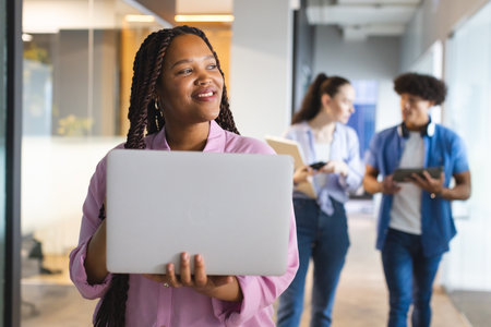 A young biracial woman holding a laptop, colleagues chatting behind in a modern business office. She...
