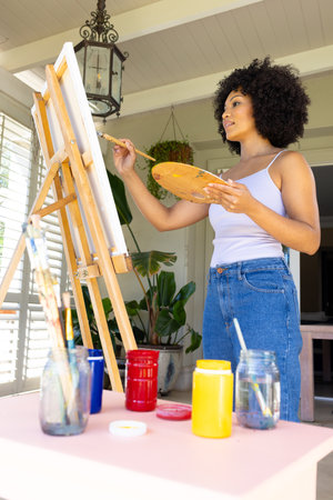 Young biracial woman painting on canvas at home, wearing casual clothes. She has curly black hair, holding palette and brush, unalteredの写真素材