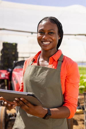 African American young female farm supervisor in greenhouse, holding tablet, smiling at camera. She has dark skin, braided hair, and wears an orange shirt with a green apron.の写真素材