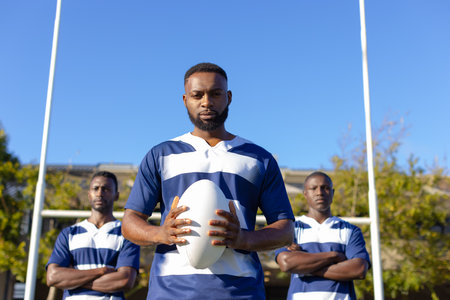 Three African American young male athletes holding a rugby ball, standing on field outdoors. All have short hair, wearing matching striped shirts and shorts, unaltered.の写真素材