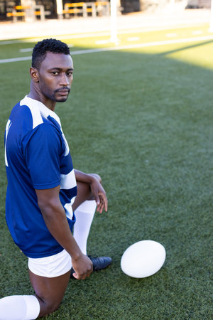 African American young male athlete kneeling on field outdoors, holding a rugby ball, copy space. He has short black hair, focused eyes, and wears a blue and white sports kit, unaltered.の写真素材