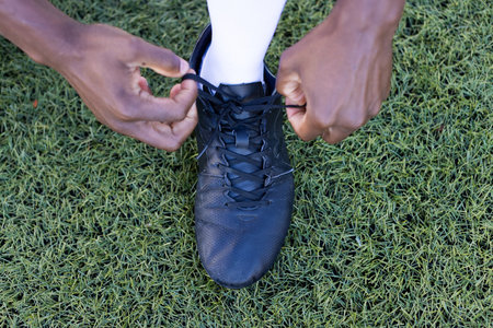 African American young male athlete tying black shoe laces on a grass field outdoors. He has dark skin and is preparing for a training session, unaltered.の写真素材