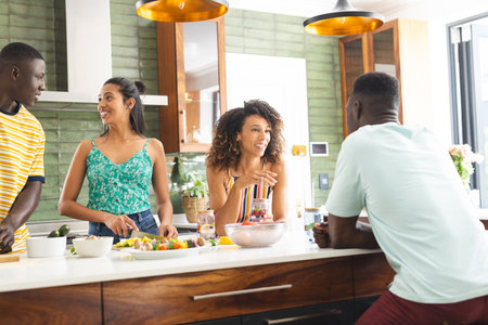 A diverse group of friends gathers in a modern kitchen. The African American male and Hispanic females chat while preparing a meal, showcasing casual attire and lively expressions, unalteredの写真素材