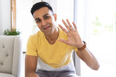 Middle Eastern man greets warmly from a bright home setting during a video call. His friendly wave adds a personal touch to the inviting atmosphere.の写真素材