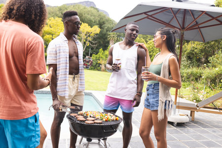 Diverse friends enjoy a barbecue outdoors, with a young African American man at the grill. They are...