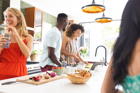 Young African American man and biracial woman prepare food together in a modern kitchen. Friends enjoy a casual gathering, with a young Caucasian woman sipping a drink, smiling.の写真素材