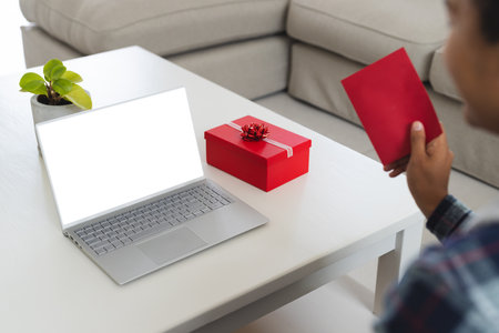 Young biracial man holding a red envelope at home, with copy space. He's at a tidy workspace with a laptop and a gift box, preparing a surprise.の写真素材