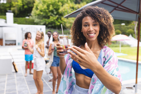Young biracial woman smiling, holding a drink at a pool party. Friends gather in the background,...