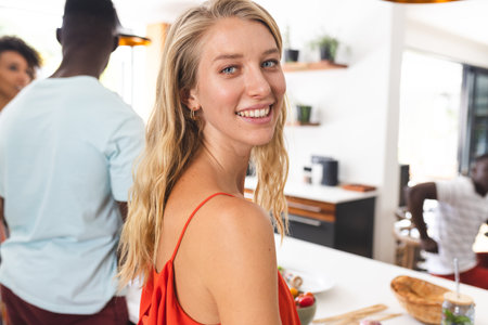 Young Caucasian woman in a red top smiles at a social gathering. Diverse friends engage in conversation in a bright, modern kitchen setting.の写真素材