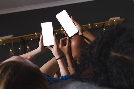 Happy diverse female friends lying on bed and using smartphones with copy space at home. Domestic life, friendship, technology and lifestyle, unaltered.の写真素材