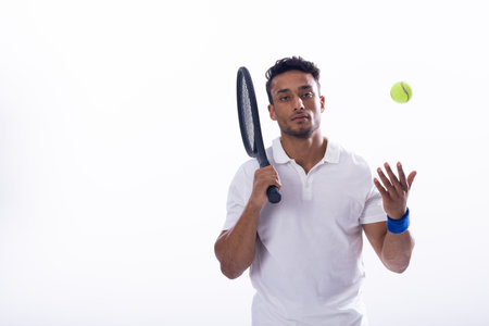 Young biracial man poised with a tennis racket and ball on white background, with copy space. He's ready for a match, showcasing skill and concentration in a studio setting.の写真素材