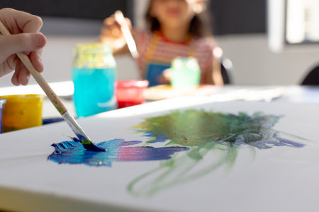 In school art class, biracial girl and a Caucasian girl, both young, painting together. Both wearing...
