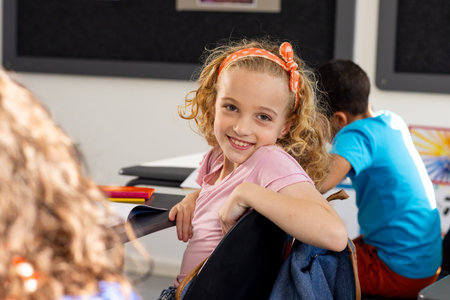 In school, young Caucasian girl with curly hair is smiling, sitting at a desk in a classroom. She has light brown hair with an orange hairband and wears a pink shirt, unaltered.の写真素材