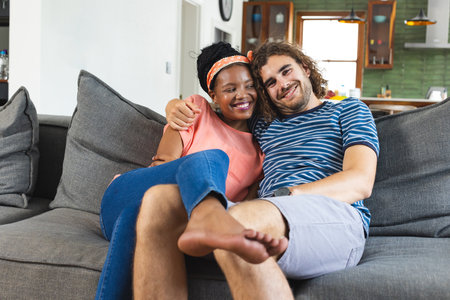 A diverse couple is sharing a cozy moment on a sofa at home. She is African American with a bright smile, headband, and orange top, while he has curly hair and a striped shirt, unaltered.の写真素材