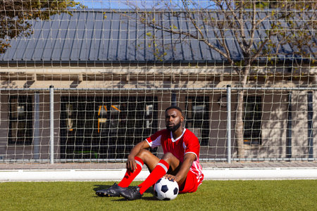 African American young male athlete resting in front of soccer goal, copy space. Wearing red sportswear, looking thoughtful, sitting outdoors, unalteredの写真素材