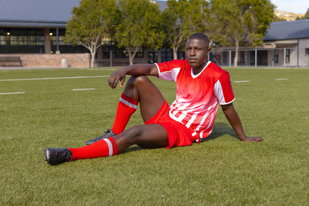 African American young male athlete sitting on grass on field outdoors, wearing red soccer uniform. He has short black hair, looking focused, with a strong and fit build, unaltered.の写真素材
