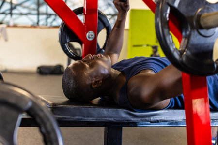 African American young male athlete in gym lifting weights, focusing on exercise. He has short hair, wearing a blue tank top, training his chest with a barbell, unaltered.の写真素材