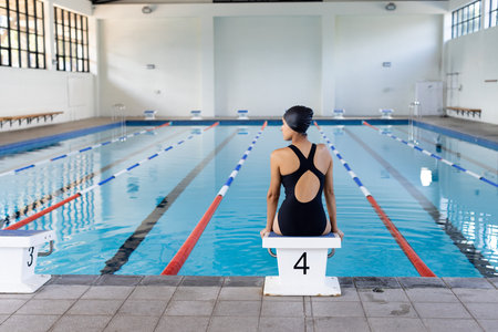 Biracial young female swimmer sitting at pool edge indoors, looking at water, copy space. She has dark hair, wearing a black swimsuit, ready to train, unalteredの写真素材